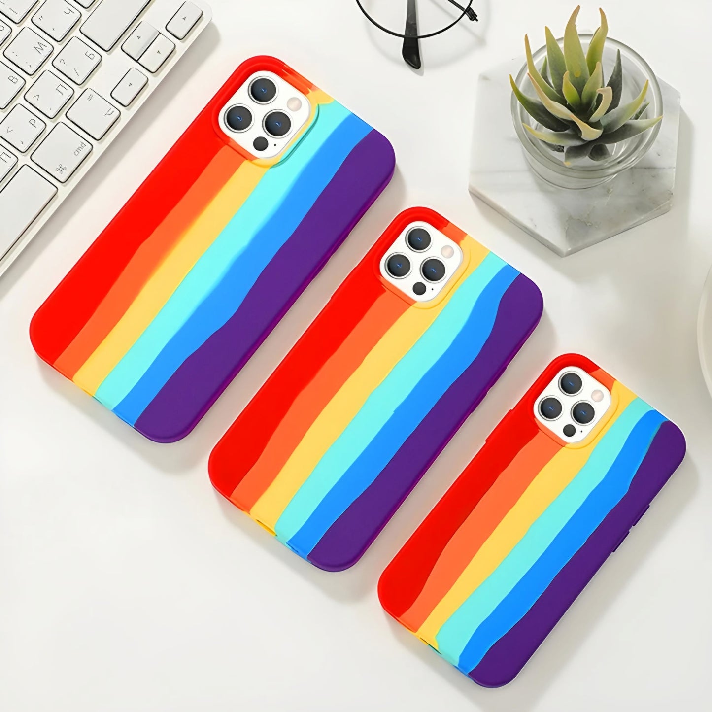 Three iPhones on a white surface, each adorned with ZIVIKART's Silicone Case for iPhone 12 Series, featuring rainbow-striped back covers in colors red, orange, yellow, green, blue, and purple. The cases boast a soft microfiber lining and are compatible with wireless charging. Beside the iPhones are a white keyboard, a pair of glasses, and a small plant in a hexagonal pot.